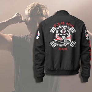 Może przedstawiać: Czarna kurtka bomberka z logo Cobra Kai na plecach. Logo przedstawia węża kobry z koreańskim tekstem i białymi paskami. Kurtka ma również naszywkę z flagą koreańską na rękawie.