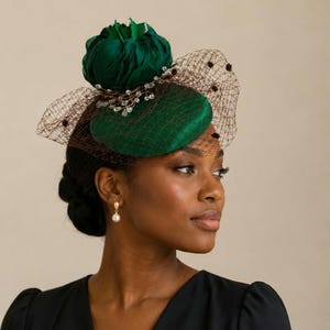 May include: A dark green fascinator hat featuring a rounded base, a large fabric flower accent, and a brown netting veil with small dark spots. The design includes delicate wire embellishments with clear beads attached to the base of the flower.