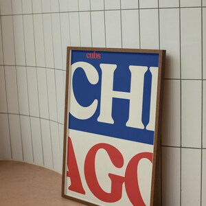 Può includere: Un poster incorniciato con la parola "CHICAGO" in grandi lettere bianche e rosse. La parte superiore del poster ha la parola "cubs" in rosso su sfondo blu. La cornice è di colore marrone scuro.