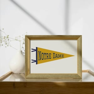 Könnte beinhalten: Ein gerahmter Druck mit einem gelben Wimpel und dem Text "NOTRE DAME" in Marineblau. Der Wimpel befindet sich vor einem weißen Hintergrund in einem goldfarbenen Rahmen. Eine weiße Vase mit Blumen ist sichtbar.