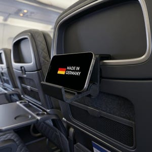Peut inclure: Un support de smartphone en plastique noir fixé au dossier d'un siège d'avion. Le support maintient un smartphone affichant le drapeau allemand et le texte MADE IN GERMANY. Le siège présente un appui-tête matelassé gris foncé et une tablette.