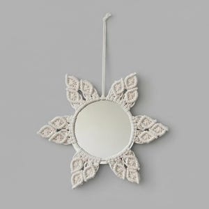 May include: A round mirror with a cream-colored macrame sunburst design. The mirror is suspended by a matching cord. The sunburst has six leaf-shaped elements surrounding the circular mirror. The mirror is a decorative wall hanging.