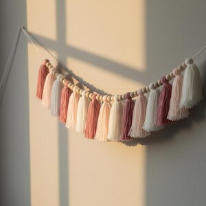 May include: A decorative tassel garland featuring a variety of pink and white tassels. The tassels are strung together with wooden beads and a white cord. The garland is hanging against a neutral wall, with sunlight casting shadows.