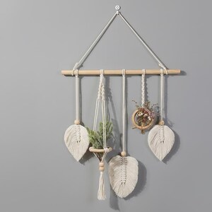 May include: Macrame wall hanging with a natural wood dowel and white cotton cord. Features three leaf-shaped accents, a small hanging planter with greenery, and a wooden hoop with dried flowers. A decorative piece for home decor.