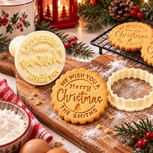 May include: A white plastic cookie stamp and a matching scalloped cutter, alongside a round golden-brown cookie embossed with the text WE WISH YOU A Merry Christmas THE SMITHS and a small pine tree icon, all arranged on a wooden board.