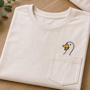 Può includere: T-shirt color crema con tasca con ricamo di un'anatra dei cartoni animati. L'anatra ha un becco giallo e sbuca dalla tasca. La maglietta è piegata ordinatamente su una superficie di legno.
