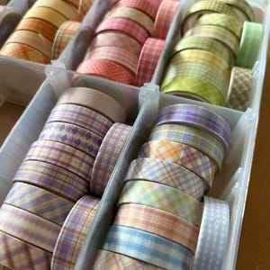 May include: A clear plastic organizer filled with rolls of decorative washi tape. The tape features various plaid patterns in pastel colors like pink, green, yellow, and purple. The rolls are neatly stacked, showcasing the variety of designs.
