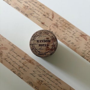 May include: A roll of light brown decorative tape featuring a vintage-style script pattern with cursive handwriting, postal stamps, and the number 32. The circular top of the roll displays the text RANDOM NOTES surrounded by the phrase ABOUT THE PAST.