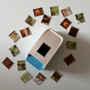 May include: A white and light blue craft punch tool with a rectangular base, surrounded by numerous small, square paper cutouts with serrated edges. Each paper cutout features a unique printed image of various outdoor and indoor scenes.