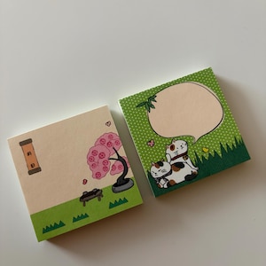 May include: Two square sticky notes with Japanese-inspired illustrations. One shows a pink flowering tree and a small bench. The other has two cats and a speech bubble. Both have green grass borders and are on a white surface.