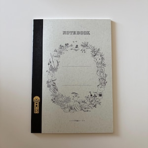 May include: A rectangular notebook with a light grey speckled cover and a black spine. The cover features the text NOTE BOOK at the top, a central wreath illustration of Moomin characters and plants, and Japanese text at the bottom. A gold logo with H30S is on the black spine.