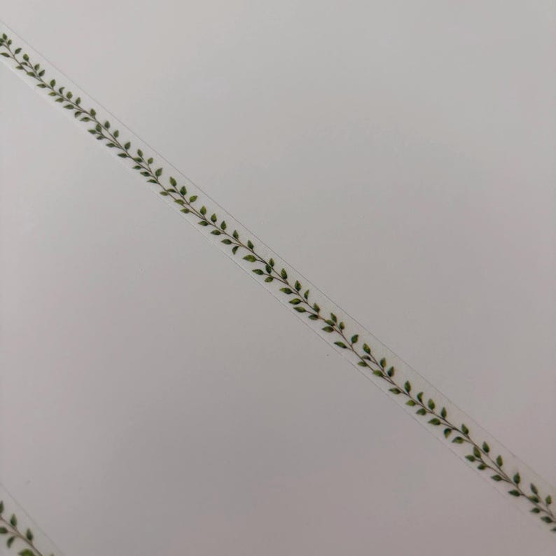 May include: A transparent tape with a repeating pattern of green leaves and stems. The tape is diagonally positioned against a white background, creating a decorative border or accent.
