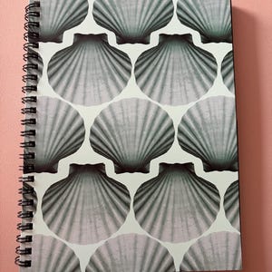 May include: A spiral-bound notebook with a repeating pattern of grey seashell designs on a white background. The cover has a light pink hue. The spiral binding is black.
