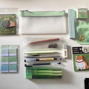 May include: A collection of stationery items including a translucent pencil case, a leaf-shaped clip, various green-toned pens, markers, sticky notes, a Milan eraser, paper clips, and a Shiba Inu-themed stand stick marker set in plastic packaging.