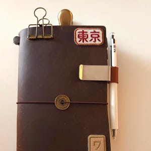 Puede incluir: Un diario de cuero marrón con un portalápices, un clip dorado y un pequeño parche que dice "Tokyo". El diario está cerrado con una banda elástica marrón y tiene una moneda decorativa. Un bolígrafo blanco está en el portalápices.