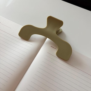 May include: A pale green hair clip sits on an open notebook. The clip has a curved design and a spring-loaded mechanism. The notebook has white, lined pages and the words "pieces of" printed at the top. The notebook's pages have thin, horizontal lines.