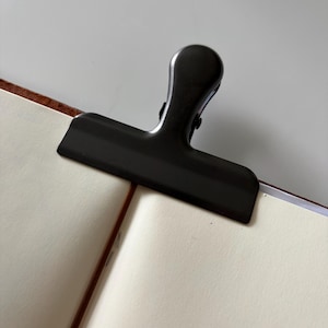 May include: A black metal binder clip securing the pages of a notebook. The clip is on cream-coloured paper, with a brown spine visible. The background is a plain white surface.