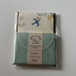 May include: A stationery set in clear packaging containing five light green envelopes and ten cream-coloured sheets of paper. The paper features a blue bird illustration with yellow and green floral accents. The label reads Fleurs et chance.