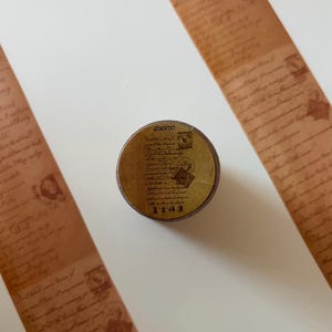 May include: A roll of decorative tape with a vintage aesthetic. The tape has a brown background with printed script and illustrations. The tape roll has the word "zoomo" printed on it, along with other text and illustrations.