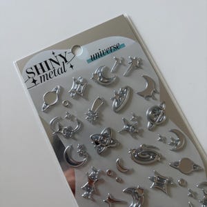 Peut inclure: Une feuille d'autocollants métalliques argentés étiquetée SHINY metal universe, présentant diverses formes célestes dont des croissants de lune, des planètes avec anneaux, des étoiles filantes et des étoiles à plusieurs branches sur un fond gris clair.