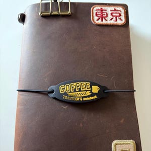 May include: A brown leather Traveler's notebook with a black elastic closure. The notebook features a gold-coloured clip, a patch with Japanese text, and a black tag that reads "COFFEE and Traveler's notebook" in gold lettering.