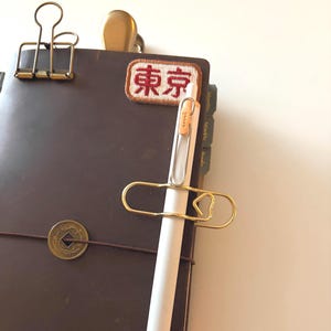 Puede incluir: Un diario de cuero marrón con un clip dorado que sujeta un bolígrafo blanco. Un parche con los caracteres japoneses de "Tokio" está adherido al diario. También se ven un clip dorado y un cierre de latón.