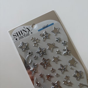 May include: A sheet of silver-coloured metallic star stickers labelled SHINY metal star. The sheet contains various sizes of star shapes, including solid stars, outlined stars, and shooting star designs with curved trails, all arranged on a grey backing.