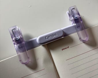 May include: A light purple plastic page holder with two translucent clips and a central bar embossed with the word Campus. The device is positioned across the open pages of a notebook featuring lined paper and the printed text pieces of.
