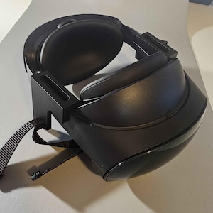May include: A black VR headset with a strap and a stand. The headset has a black headband and a black faceplate.