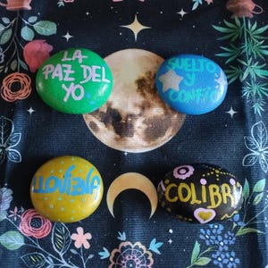 Könnte beinhalten: Vier bemalte Steine mit inspirierenden Botschaften. Die Steine sind grün, blau, gelb und schwarz. Der grüne Stein sagt "LA PAZ DEL YO", der blaue Stein sagt "SUELTO Y CONFIO", der gelbe Stein sagt "LLOVENA" und der schwarze Stein sagt "COLIBRI".