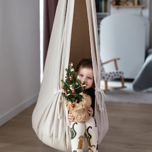Puede incluir: Un niño está sentado en una silla colgante de lona beige, sosteniendo un pequeño árbol de Navidad decorado en una maceta de papel. El niño lleva leggings estampados y una camisa negra. La silla está suspendida del techo.