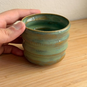 Könnte beinhalten: Eine handgefertigte Keramiktasse mit einer strukturierten, geriffelten Außenseite. Die Tasse hat eine Mischung aus grüner und brauner Glasur, die eine rustikale, erdige Ästhetik erzeugt. Der Innenraum ist ein glänzendes, tiefes Grün. Die Tasse wird von einer Hand gehalten.