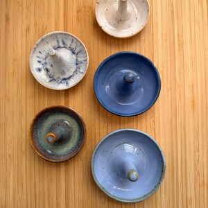 Op de afbeelding: Vijf handgemaakte keramische ringhouders in verschillende kleuren, waaronder blauw, wit en bruin. Elke houder heeft een kleine, verhoogde paal in het midden om ringen vast te houden. De ringschalen zijn gerangschikt op een lichtbruin houten oppervlak.