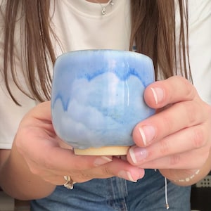 Può includere: Una tazza in ceramica fatta a mano con una glassa azzurra. La tazza ha una forma arrotondata e una base color crema. La glassa blu ha un effetto nuvoloso, acquerello. La tazza è tenuta in due mani.