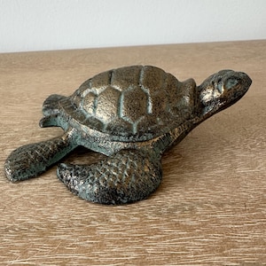 May include: A bronze-colored turtle figurine with a textured shell and flippers. The sculpture has a metallic finish with hints of teal and is resting on a wooden surface. The turtle is a decorative item.