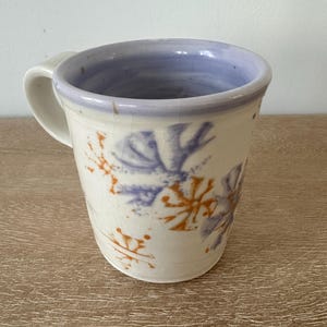 May include: A handmade ceramic mug with a white exterior, a light purple interior, and a white handle. The mug is decorated with blue and orange floral patterns. The mug is sitting on a wooden surface.