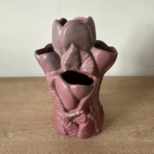 May include: A dusty rose ceramic vase featuring a sculptural design of multiple overlapping tulip-shaped openings. The surface has a matte, slightly speckled texture with subtle variations in color depth across the molded floral forms.