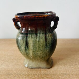 May include: A ceramic vase featuring a two-toned drip glaze finish. The upper portion is a deep, glossy brown, while the lower body transitions into a textured, mossy green and cream color. It has two small, integrated handles on the sides.
