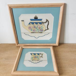 May include: Two framed cross-stitch artworks featuring a teapot and a teacup. Each piece displays a cream-colored vessel with blue wave patterns and floral accents, resting on a white doily, set within light wood frames and blue matting.