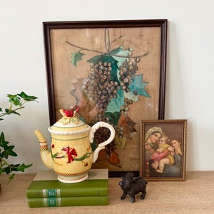 May include: A yellow ceramic teapot with a red bird finial, floral accents, and a checkered base, resting on two green books. Behind it are two framed pictures: one depicting grapes and holly leaves, and the other a portrait of a person holding a child.