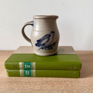 May include: A beige stoneware pitcher with a blue hand-painted bird design on the front, featuring a curved handle and a wide rim. The pitcher is resting on two stacked green hardcover books titled Jack and Jill and Little Men by Louisa May Alcott.
