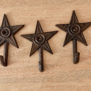 May include: Three dark brown cast iron wall hooks, each featuring a five-pointed star design with a central circular detail and a curved hook extending from the bottom point, designed for mounting with visible screw holes at the top.