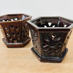 May include: Two hexagonal ceramic planters with a dark brown glossy glaze. Each planter features intricate cut-out floral patterns on all sides and includes a matching detachable hexagonal saucer base for drainage and stability.