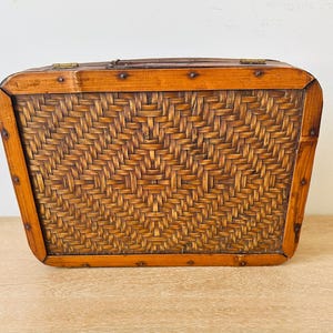 May include: A rectangular brown woven basket with a solid wooden frame. The basket features a dark brown intricate woven pattern on the front panel, secured by a smooth wooden border with visible metal nails and two brass hinges on the top edge.