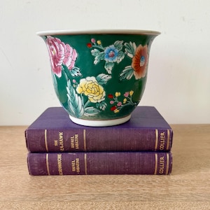 May include: A green ceramic planter featuring a hand-painted floral pattern with pink, yellow, blue, and orange blossoms and green leaves. The bowl-shaped vessel has a wide, flared rim and a white base, resting atop two stacked purple hardcover books.