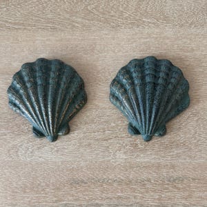 May include: Two identical scallop shell wall decorations crafted from dark metal with a weathered, bronze-toned patina finish. Each shell features a fan-shaped design with prominent vertical ridges and a textured surface, showing a realistic shell form.