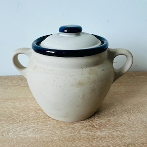May include: A beige ceramic pot with two side handles and a matching lid. The pot features a dark navy blue glazed rim and a matching dark navy blue knob on the center of the lid. The body of the pot has a matte, light tan, textured finish.