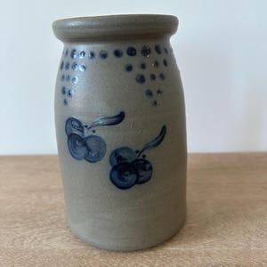 May include: A stoneware jar with a gray-blue glaze. The jar features dark blue floral designs and a pattern of dots near the rim. The jar has a slightly tapered shape and a rounded top. A rustic, handmade aesthetic.