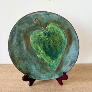 May include: A round ceramic plate featuring a prominent embossed green leaf design at the center. The plate has a mottled blue and brown glaze finish with a textured surface. It is displayed on a dark red stand, showcasing the detailed leaf veins.
