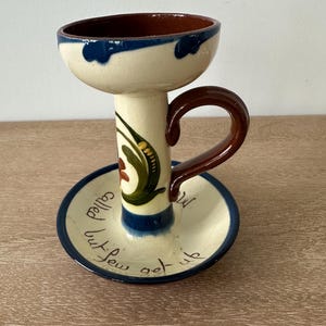 May include: Hand-painted ceramic candle holder with a cream base, blue trim, and a brown handle. The candle holder has a circular base with the words "called but few get up" written on it. The design includes green and red floral accents.